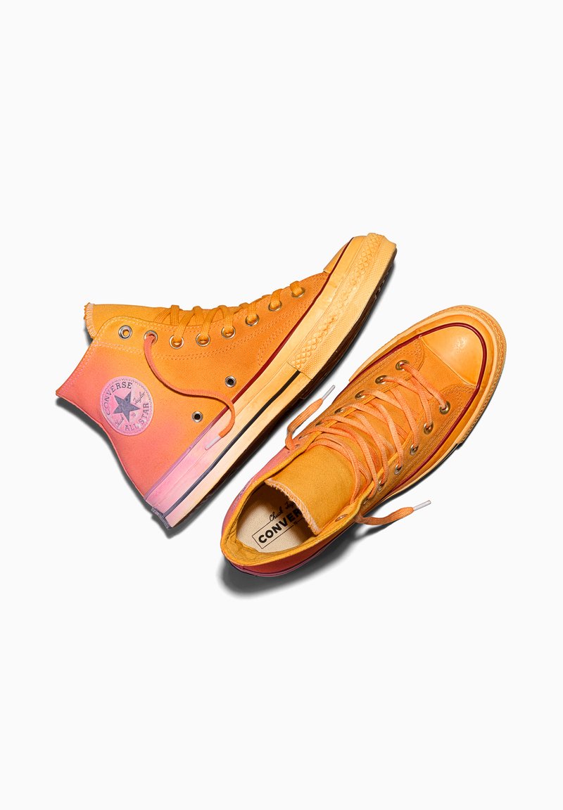 Paar orangefarbene High-Top Converse-Sneaker mit Schnürsenkeln, Gummisohlen und Logo-Patch am äußeren Knöchel vor weißem Hintergrund.