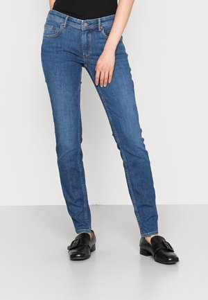 Jeans de mezclilla azul con un corte slim, diseño de cinco bolsillos, costuras sutiles y un cierre de botón y cremallera. Llevados con mocasines negros.