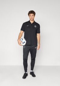 Sort polo med logo, matchende sorte træningsbukser og sorte fodboldstøvler. Model holder en hvid og blå fodbold.