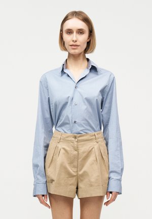 Γυναίκα με κοντά ξανθά μαλλιά που φοράει ανοιχτό μπλε ριγωτό πουκάμισο με κουμπιά και μπεζ βερμούδα με πιέτες και ψηλή μέση, στέκεται μπροστά σε λευκό φόντο.