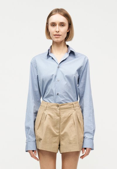 Femme aux cheveux blonds courts portant une chemise rayée bleu clair à boutons et un short beige taille haute à plis, debout devant un fond blanc.