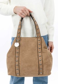 Sac à main en suède beige avec des bordures noires, présentant de grands clous en métal argenté sur les côtés et une étiquette circulaire attachée à la poignée.