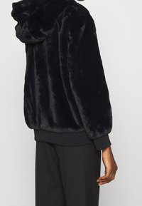 Zwarte nepbont hoodie met een zachte textuur, verlaagde schouders en geribde manchetten. Heeft een capuchon en een relaxte pasvorm.