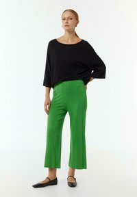 Schwarzes, locker sitzendes Oberteil und knielange, leuchtend grüne, gerippte, weite Hose. Schwarze flache Schuhe runden das Outfit ab. Heller Hintergrund.