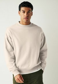 Sweatshirt beige clair avec un col rond côtelé, coupe surdimensionnée et manches à poignets, associé à un short vert foncé, texture douce.