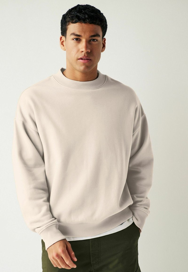 Sweatshirt beige clair avec un col rond côtelé, coupe surdimensionnée et manches à poignets, associé à un short vert foncé, texture douce.