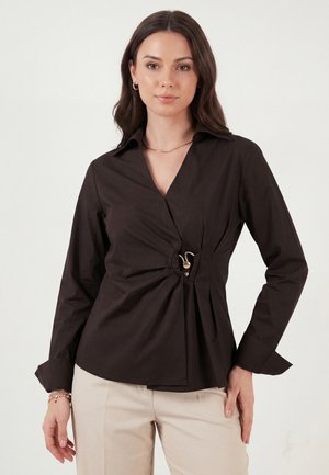 Kobieta w czarnej bluzce z długim rękawem z centralnym marszczonym detalem i beżowych spodniach, stojąca na jednolitym białym tle.