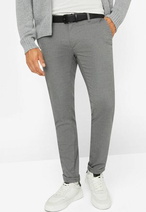 Mann trägt graue, strukturierte, schmal geschnittene Hose, schwarzen Gürtel, weiße Sneaker und grauen Cardigan mit den Händen in den Taschen.