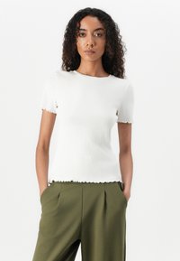T-shirt bianco a coste con maniche corte e bordi ondulati, abbinato a pantaloni verde oliva a vita alta. Tessuto liscio e strutturato.