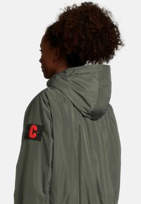 Olijfgroene hoodiejack met een gladde textuur, een zwarte stoffen patch met een rode letter 'C' op de linkermouw, en een strakke nek.