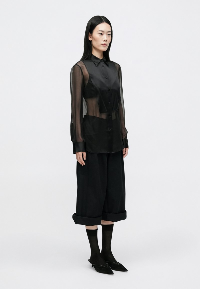 Chemise noire transparente à manches longues, associée à un pantalon large noir, des chaussettes noires transparentes et des chaussures noires à bout pointu.