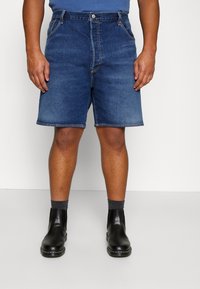 Shorts de mezclilla en azul medio con corte recto, cinco bolsillos y cremallera. Combinados con botas de cuero negras y calcetines grises.