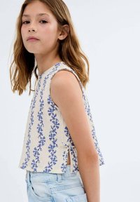 Top court sans manches en crème avec un motif floral bleu ; présente un col rond et des fentes sur les côtés, fabriqué dans un tissu léger.