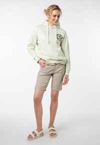 Lichtgroene hoodie met trekkoorden, beige shorts en witte sandalen. De hoodie heeft een graphic op de borst en een ontspannen pasvorm.