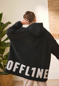 Czarna, oversize'owa kurtka polarowa z kapturem. Posiada odważne białe napisy "OFFL1NE" u dołu. Miękki, teksturowany materiał.