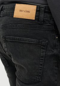Οπίσθια όψη μαύρων τζιν με μεγάλη πίσω τσέπη και μπρονζέ δερμάτινη ετικέτα της μάρκας "ONLY & SONS" στη ζώνη μέσης.