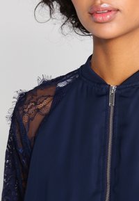 Tmavě modrá bunda s krajkovými rukávy a zapínáním na zip. Obsahuje strukturované krajkové detaily na ramenou a kulatý výstřih.