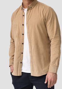 Beige Cord-Hemd mit Knopfkragen, ausgestattet mit dunklen Knöpfen, geradem Schnitt und weicher Textur, über einem weißen Hemd getragen.