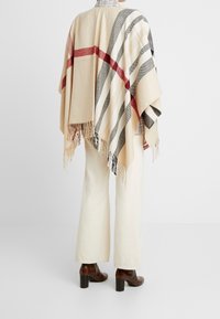 Personne portant un pantalon large crème, des bottes à talons marron et un poncho beige avec des motifs à rayures rouges et noires ainsi que des détails en franges.