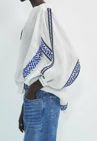Witte linnen blouse met blauwe geborduurde patronen langs de mouwen, met een losse pasvorm en geplooide details. Gecombineerd met blauwe spijkerbroek.