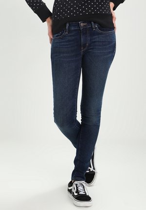 Jeans ajustados de denim azul oscuro, con un clásico diseño de cinco bolsillos y un ligero desvanecimiento. Combinados con zapatillas negras.