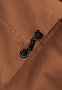 Bruine stof met een gladde textuur, voorzien van zwarte hardware en een trekkoordsluiting aan een rand, met opvallende stiksels.