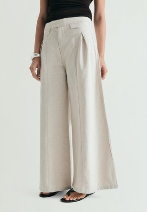 Femeie purtând pantaloni bej cu croială largă, top negru fără mâneci, brățară argintie și sandale negre, stând în picioare în fața unui fundal simplu, deschis.