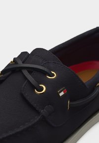 Μαύρο δερμάτινο παπούτσι τύπου boat με χρυσά ρουθούνια, μαύρα κορδόνια και ένα μικρό ορθογώνιο λογότυπο σε κόκκινο, λευκό και μπλε στη πλευρά.