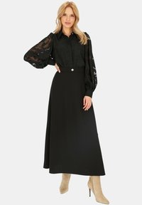 Czarna bluzka z przezroczystymi, wzorzystymi rękawami, w połączeniu z długą czarną spódnicą o dopasowanej talii i metalicznym akcentem. Beżowe kozaki na obcasie.