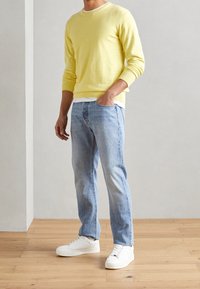 Gelbes Rundhals-Pullover, hellblaue Jeans und weiße Sneakers. Der Pullover hat gerippte Ärmelbündchen und Saum, und die Jeans sind leicht tailliert.