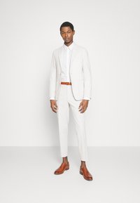 Helles Anzug mit texturiertem Finish, kombiniert mit einem weißen Hemd und braunen Lederschuhen. Verfügt über einen maßgeschneiderten Schnitt und einen braunen Gürtel.