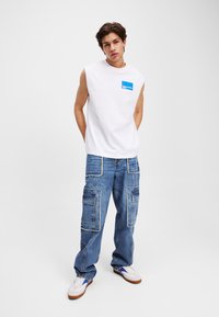 Tricou alb fără mâneci cu emblemă albastră, asortat cu pantaloni cargo din denim albastru, cu multiple buzunare și margini desfăcute. Adidași albi.