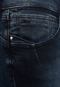 Poche de jean en denim bleu foncé avec des rivets argentés et une petite étiquette de marque en métal noir sur la partie supérieure de la poche.