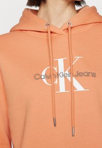 Peach-colored cotton hoodie featuring a white Calvin Klein Jeans logo, drawstrings with metal tips, and a relaxed fit.