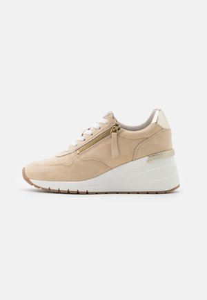 Beżowy sneakers na koturnie z białą podeszwą, białymi sznurowadłami, bocznym zamkiem błyskawicznym i metalicznym akcentem na pięcie, pokazany z lewego profilu na białym tle.