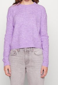 Lila gerippter Strickpullover mit Rundhalsausschnitt und langen Ärmeln, kombiniert mit hellgrauen Jeans aus Denim, die Taschen und dezente Ausfransungen zeigen.