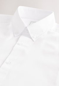 Chemise blanche boutonnée avec col pointu et boutons blancs visibles, présentée à plat sur un fond clair.