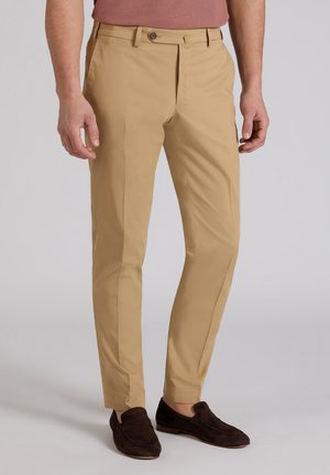 Mężczyzna w beżowych, dopasowanych spodniach i ciemnobrązowych zamszowych mokasynach, stojący na tle prostego jasnoszarego tła.