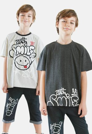 Zwei Jungen tragen grafische T-Shirts: eines in Grau mit schwarzem und rotem Design und ein anderes in Dunkelgrau, beide mit dem Aufdruck "SMILE" in Weiß. Sie tragen Jeanshosen mit umgeschlagenen Säumen.