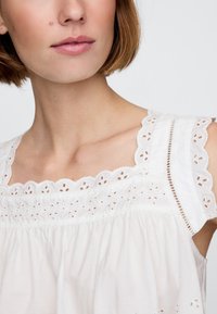 Camicia bianca di cotone con scollo smerlato e dettagli floreali ricamati sulle spalline, con una silhouette ampia e fluente.