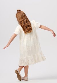 Witte geborduurde jurk met pofmouwen en kantafwerking, met bloemmotieven en een vloeiende silhouet, gecombineerd met lichtgekleurde schoenen.