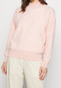 Nainen, joka pukeutuu vaaleanpunaiseen pitkähihaiseen neuleeseen ja beigeihin housuihin, seisoo yksinkertaista taustaa vasten.