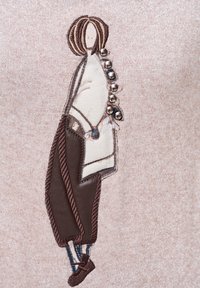 Wyhaftowana tkanina przedstawiająca stylizowaną postać z brązowymi włosami, ubrana w biały top i brązowe spodnie z dekoracyjnymi guzikami i dodatkami.