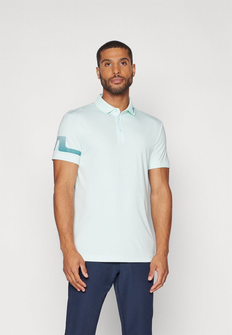 Hellblaues Poloshirt aus glattem Stoff, mit Kragen, einer Knopfleiste mit drei Knöpfen und einem strukturierten Muster auf dem linken Ärmel.