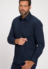 Camicia a maniche lunghe blu navy con colletto a bottone, taschino sul petto e texture liscia. Caratterizzata da bottoni scuri e design aderente.