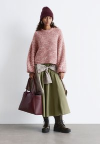 Rosa strukturierter Pullover, olivgrüne Midi-Rock mit einer Schleifendetail, burgunderfarbene Handtasche und schwarze Schnürstiefel. Die Accessoires umfassen Creolen und eine Mütze.