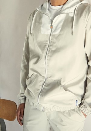 Personne portant une veste à capuche zippée beige clair avec des poches avant et un pantalon beige assorti, debout à côté d'une chaise en bois.