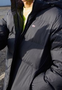 Sort pufferjakke med en kviltet design, der har høj krave, lynlås lukning og et lille logo på brystet. Tekstureret materiale.