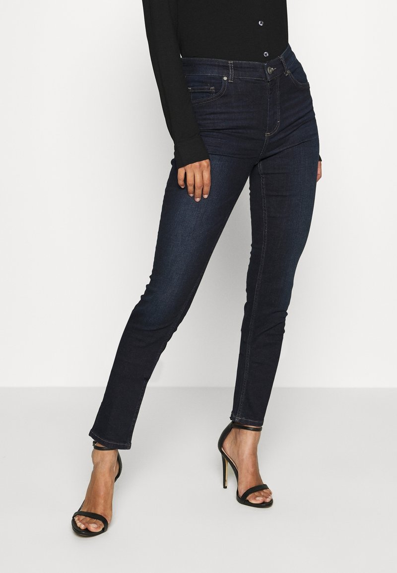 Jeans skinny en denim foncé avec une taille mi-haute, des détails de couture subtils et une silhouette ajustée, accompagnés de talons hauts noirs à lanières.
