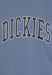 Mėlynas medvilninis marškinėlis su žodžiu "DICKIES" ryškiomis juodomis raidėmis, apsuptomis baltu kontūru, rodomas išlenktu dizainu per krūtinę.